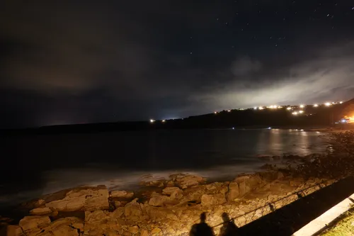 The landscape photograph shows nighttime photographers at the coast of Sennen Cove in Cornwall. Taken at night during the autumn season, the image captures the rocky shoreline under a dimly lit sky, with a faint glow from the lights of houses spread along the hillside in the distance. The shadows of the photographers can be seen in the foreground, cast by artificial lighting along the coastal path. The sky above Sennen Cove is partially cloudy, with stars visible in the clearer areas. The photograph highlights the tranquil atmosphere of the Cornish coast at night, with gentle waves creating a misty effect on the water’s surface near the rocks.